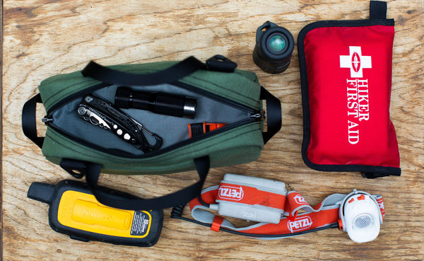 a top=down photo of a Boreal Grab Bag with camping items on a table next to it including a headlamp, a flashlight, and a First Aid Kit.